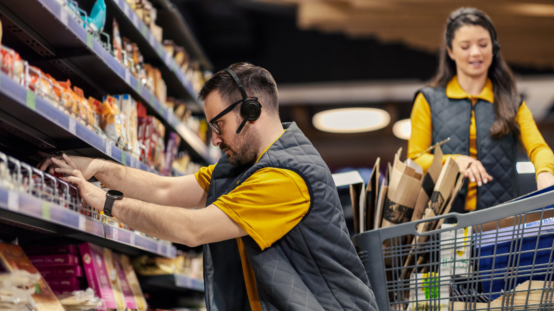 Two store employees wearing Jabra Perform 55 headsets stock shelves and move supplies through a retail aisle.